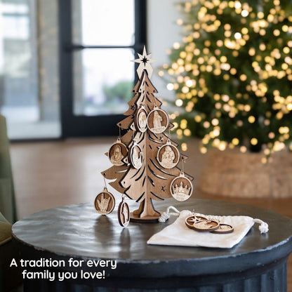 A wooden Christmas ornament tree display with carved family portrait ornaments and a star topper, set on a dark round table beside a small white drawstring pouch. A blurred Christmas tree with warm lights and an indoor room background are visible behind it.